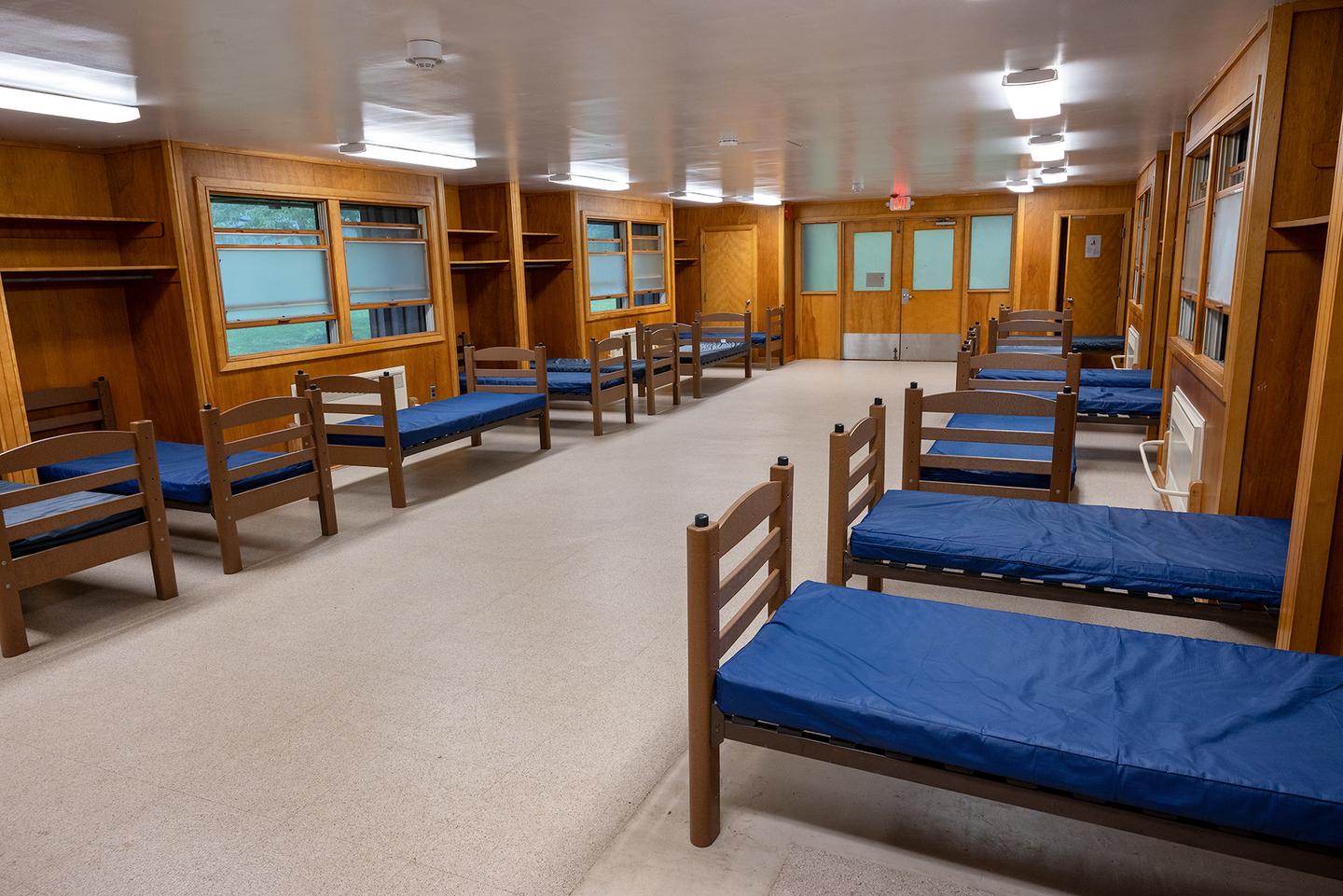 Interior view of 1 dormitory wing. Vinyl floor with wooden walls, 2 double windows, 1 double door with window panels.  10 single beds with vinyl covered mattresses.Each dormitory is heated and  accommodates 30 overnight guests.  There are 15 single beds on each of the 2 connected wings.  Each wing has a separate room with 1 single bed.  There are a set of bathrooms for each wing and a supply closet with cleaning equipment.