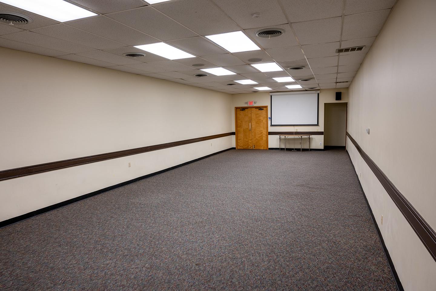 Interior view of the small conference room. Gray carpet flooring with 2 doors at the far end.  Ceiling mounted video screen on far end of room.The conference room is located inside the gym building and can accommodate 50 people. Chairs and 6 ft. tables are available in the adjacent storage room.