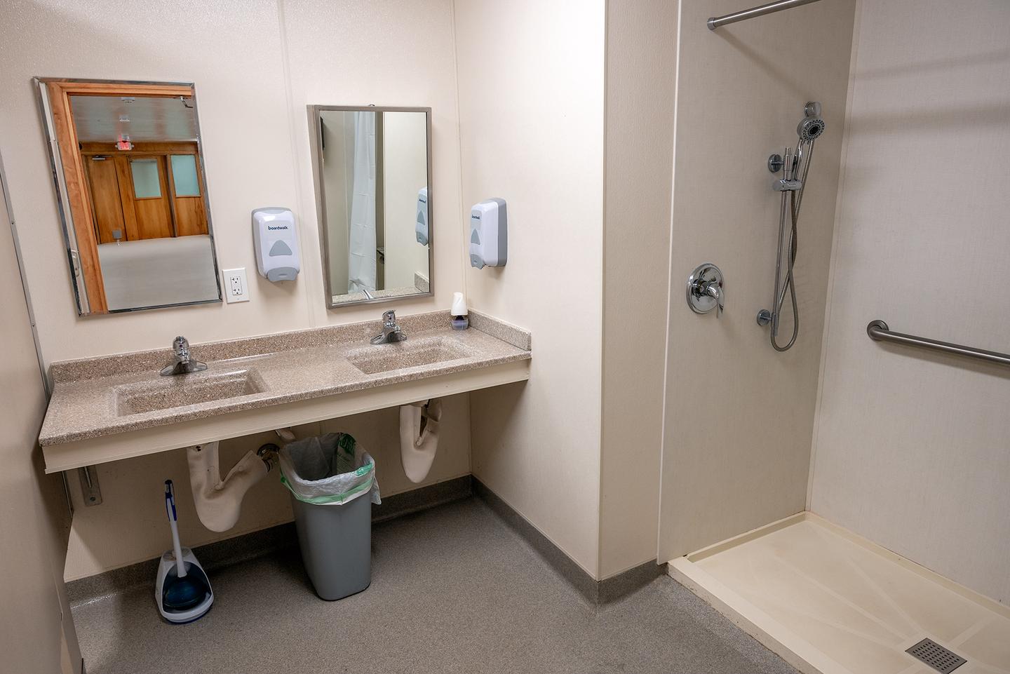 Dorm bathroom featuring one large shower, two sinks, two mirrors, two soap dispensers, and a trashcan. Each dorm has two bathrooms; one bathroom is located in each wing of the building. Each bathroom contains one shower with a folding shower chair, two sinks, and two mirrors. Not included in the photo are two toilet stalls. 