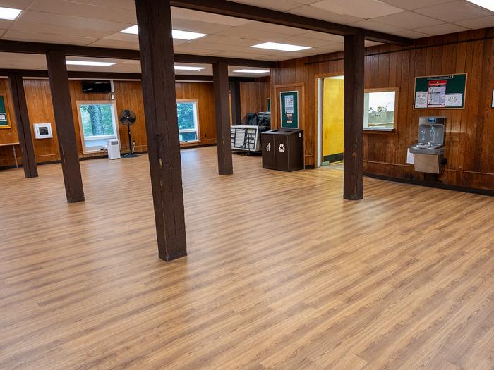 Interior view of dining hall shows 5 large wooden beams, oak laminate flooring, portable a/c unit, pedestal fan, stacked chairs, 6 ft. tables, bottle filling station, waste/recycle bin, and doorway to dishwashing room.