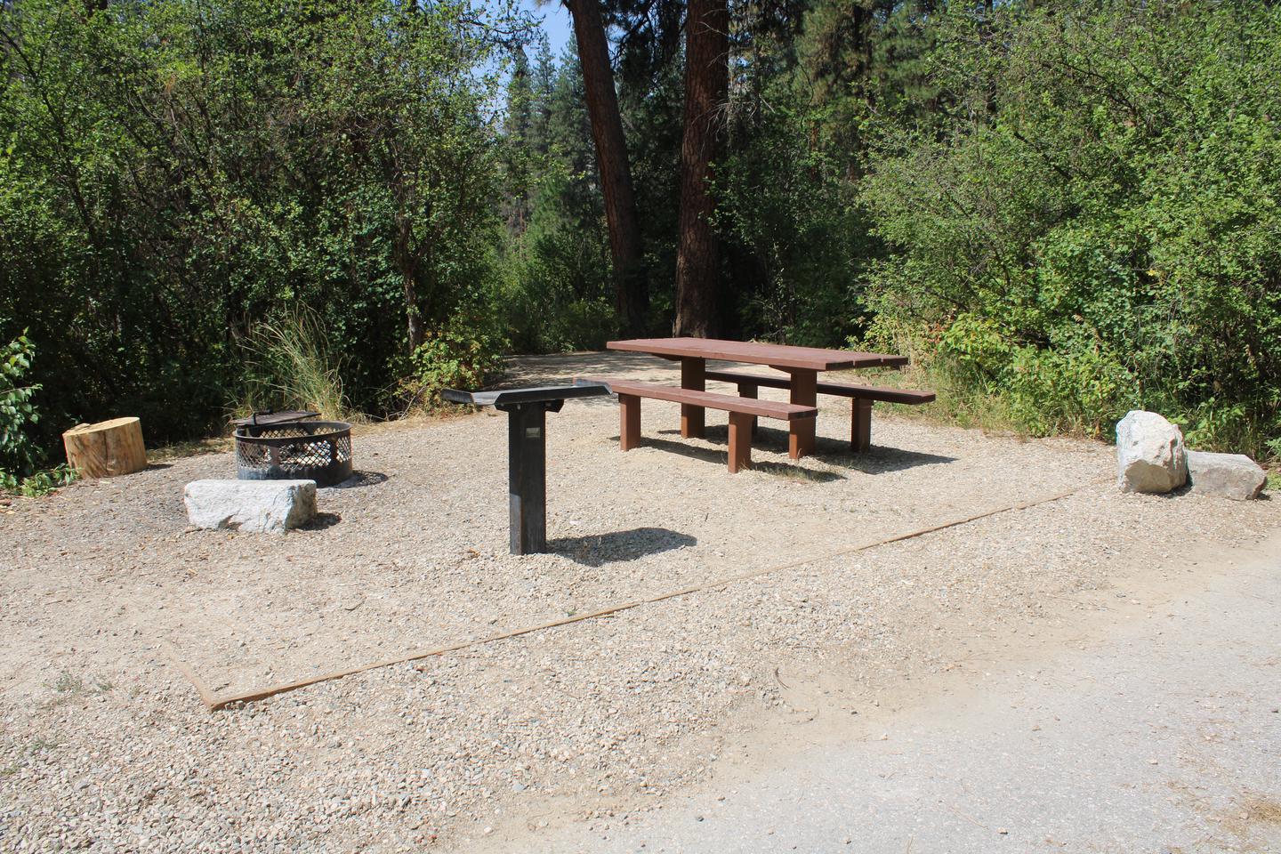 Site amenities on a sandy pad in front of trees  Site fire ring, metal table, and picnic table