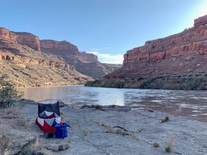 A tent pitched on a rocky ledge of the river, with towering red-rock peak buttes under a blue skyRiver camps are typically found on rocky ledges or sandbars, depending on time of year and water levels