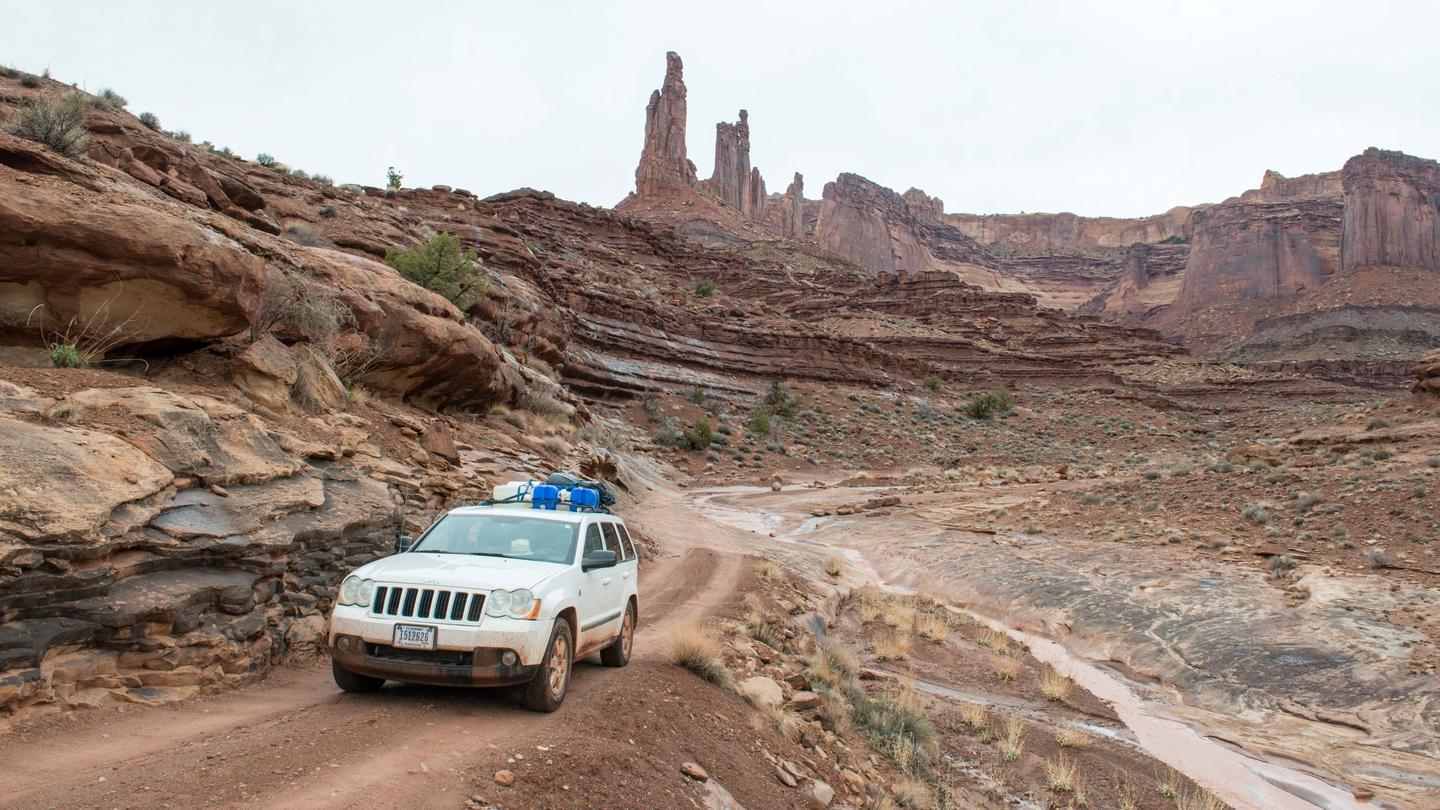 A white 4WD vehicle passes towering red-rock formations on the dirt road Driving Past Monster Tower and Washer Woman Rock Formations