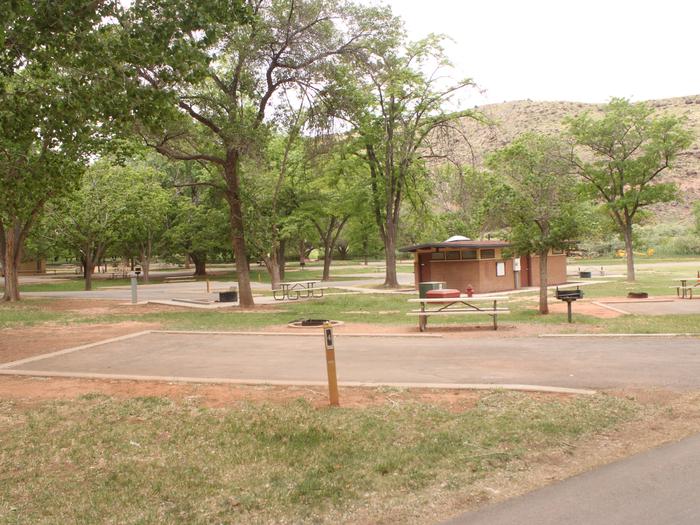 A paved driveway. Facing the end of the driveway, a picnic table and grill are off to the right side. A fire pit is behind them. A small building is in the background.Site 4, Loop A in summer.
Paved Dimensions: 14' x 36'