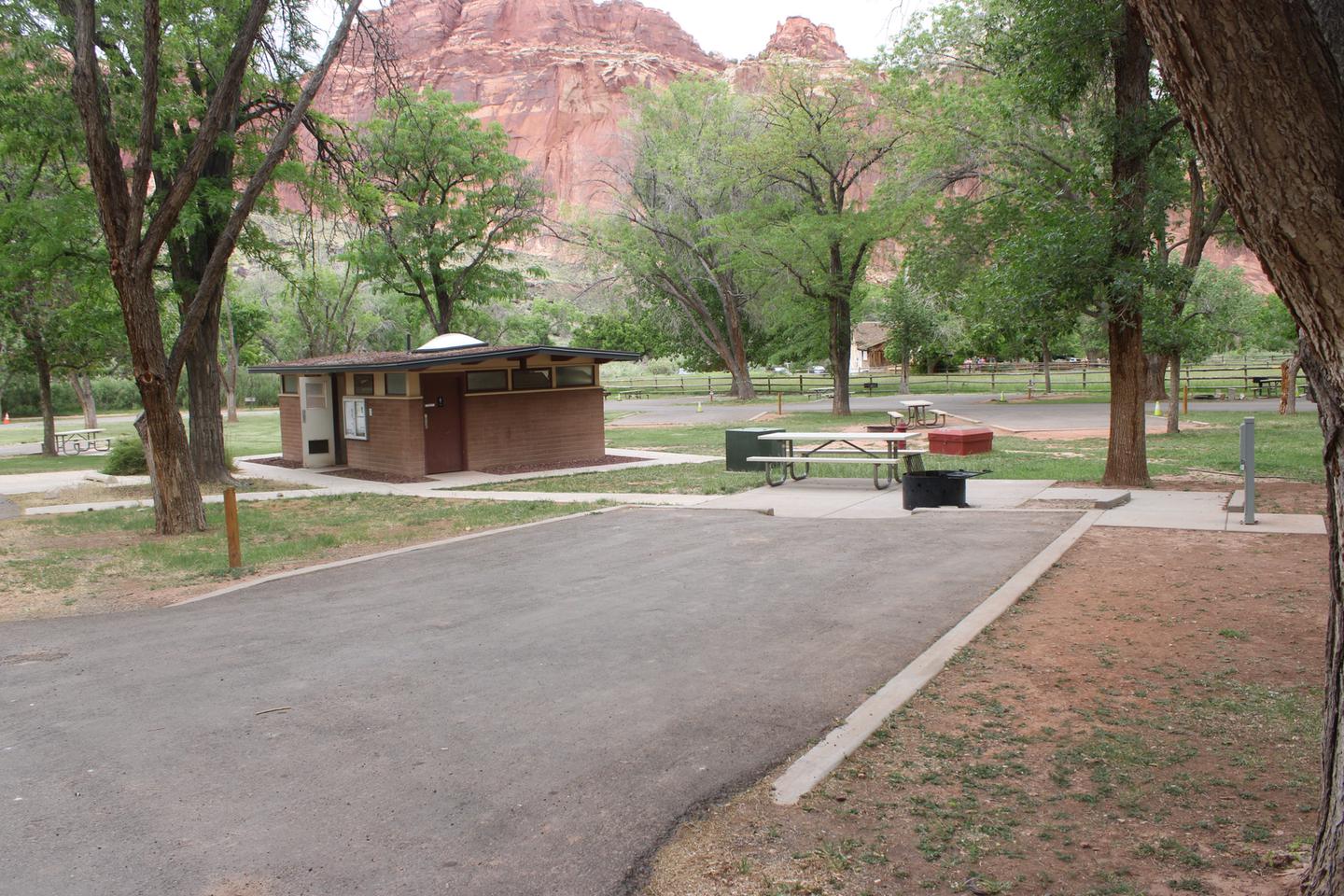 A paved driveway. A picnic table and raised fire pit is directly behind the driveway both on pavement. A tree is directly behind the fire pit. A paved sidewalk comes off of the end of the driveway on the left side (facing the end of the driveway). The sidewalk leads to a small building. A gray box is at the end of the driveway, on the right hand side (facing the end of the driveway).Site 14, Loop A in summer.  Accessible site.
Paved dimensions: 15' x 28'