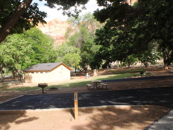 A paved driveway. Facing the end of the driveway, a picnic table, grill, and fire pit are to the left. A small building is behind the fire pit. Many trees are in the background and a red cliff rises above the trees.Site 34, Loop B
Paved Dimensions: 14' x 28'