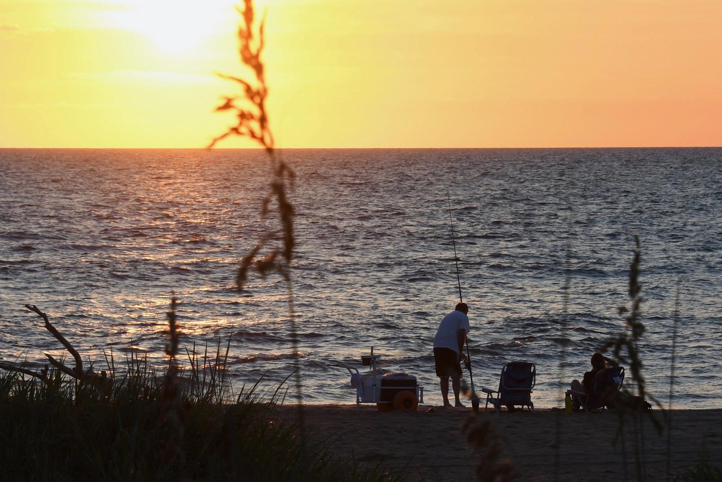 A fisherman stands on the beach with his pole. A beach chair and cooler are by his side. The sun rises over the ocean and a piece of dune vegetation covers slices through the foreground of the image.A surf fisherman enjoys an early morning on the beach at Back Bay National Wildlife Refuge in Virginia Beach, Virginia.