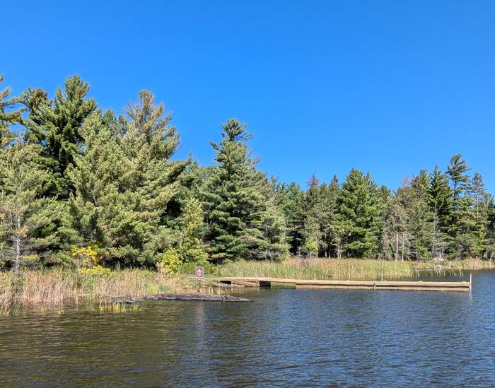 R65 - Reuter Creek, view looking at dock access from water with a campsite sign on small rock point, weedy shoreline, and towering trees in the background. View of dock boat access from the water