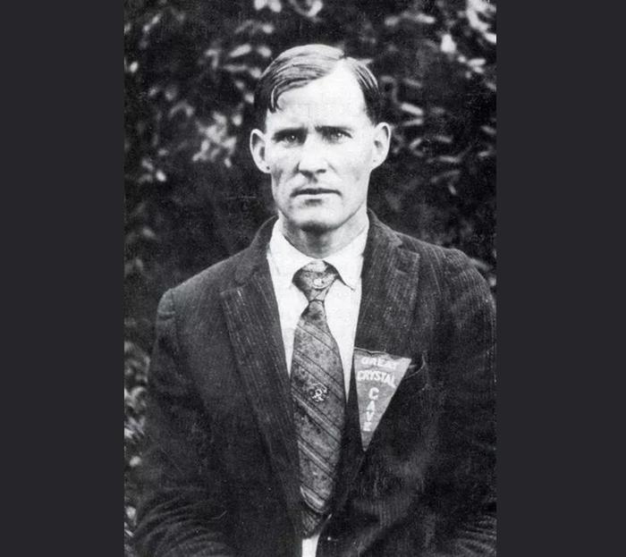 A black and white photo of a man in a pin-stripe suit. His hair is parted to the left side and his face is stoic. HIs lapel has triangular a pin reading "Great Crystal Cave." His tie has a faint pattern of diagonal lines and shifting floral patterns.  Floyd Collins
