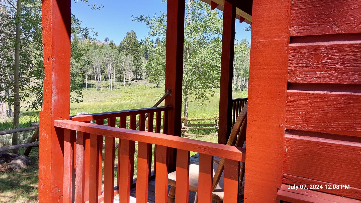 bright red wooden rails frame the front porch of the guard station where a wooden chair offers a view of the adjacent meadow on a sunny day.Front porch of Guard Station with view of adjacent meadow.