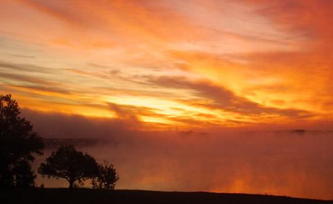 This is a picture of the sun rising across the lake. The sky is orange and yellow and there is fog rising off the lake. Sunrise photo on the Lake. 