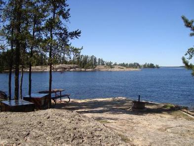 A fire ring, picnic table, and metal, bear-proof food locker on a rock outcrop, surrounded by a large, scenic lake.Looking out from a campsite onto the waters of Voyageurs National Park. 