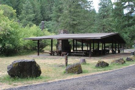 Large picnic shelter with stone fireplace and chimney and boulders lining a gravel road in front of fir forest and shrubs.WOLF CREEK GROUP SITE