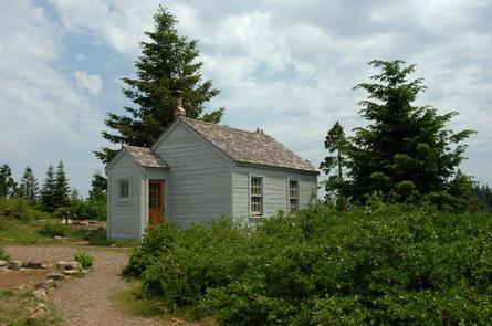 Green shrubs next to a gravel path leading to a small gray cabin with a partly cloudy sky and two tall conifer trees as a backdrop.   BUTLER BUTTE CABIN