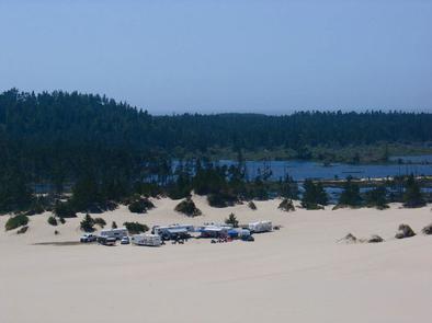 Group of trailers and trucks on a large expanse of sand near a conifer encircled lake under a blue sky.HORSFALL SAND CAMPING