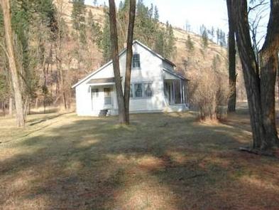 White cabin on flat ground in an open forest of conifers and deciduous trees and shrubs with no leaves.TUCANNON GUARD STATION