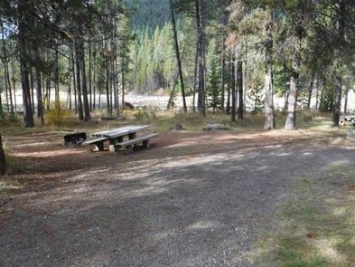 Flat gravel parking area next to picnic table and fire ring in pine forest with lake in the background.EAST LEMOLO CAMPGROUND