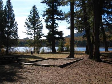 Sunlit tent pad in front of conifers in silhouette with lake and hill in background.LOST LAKE GROUP UNIT